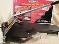 Side profile of the laminate cutter showing metal cutting mechanism, long silver handle with black grip, and black plastic base with measuring guide and orange adjustment knobs, with original packaging box in the background.