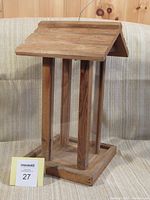 Tall wooden bird feeder with a slanted roof, clear plastic sides between wooden columns, and a base, sitting on fabric surface.