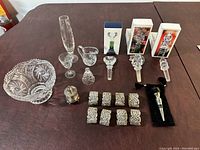 Photo of assorted crystal items including bowl, cream pitcher, bell, oblong dish; plus crystal wine stoppers in boxes, silver wine stopper with pouch, vintage salt shaker and silver-tone napkin rings arranged on brown table.