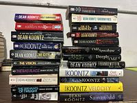 A stack of Dean Koontz novels showing a variety of titles and colors indicating a large collection of paperbacks and hardcovers in fair condition.