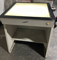 Front view of Colight light table with illuminated glass top, showing white metal frame, black frame around glass top, and open lower shelf.