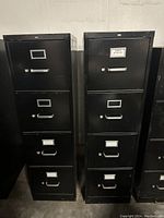Front view of three tall black metal file cabinets, showing four drawers each with handles and label holders, some visible wear.