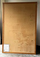 Full view of the large wood framed cork bulletin board standing upright against a wall. Shows the cork surface with multiple push pins attached and some wear marks.