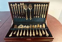 Open mahogany flatware chest showing gold-plated knives, forks, spoons, and serving utensils arranged in velvet slots.