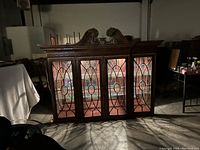 Large wooden hutch top section with ornate carved scrollwork on top and four glass doors with detailed wood muntins, lit interior visible.
