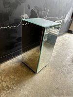 Mirrored rectangular end table standing on concrete floor, showing reflective surfaces and minor scratches visible on top edge.