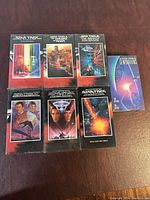 Seven Star Trek original cast movie VHS tapes displayed on a brown surface. Six tapes are still in sealed plastic wrap, one tape (Star Trek VI: The Undiscovered Country) is unwrapped.