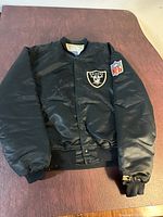 Black vintage Oakland Raiders jacket with NFL patch and visible stain on the front, presented flat to show condition and details.