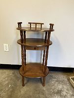 Full front view of small vintage wooden table with three tiers, including top with decorative spindles, middle with small drawer, and bottom shelf; table stands against light wall on concrete floor.