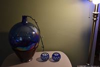 Blue glass bottle on mahogany base with two small blue decorative glass bowls on side, lit by a standing lamp, showing sheen and reflections on bottle surface