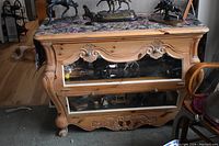 Front side view of blond wood French style display cabinet with glass shelves and decorative carvings, showing floral fabric on top and horse figurines inside the glass shelves.