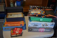 Educational games and activity kits including Fractiles-7 magnetic puzzle, origami airplane, bubble machine, dominoes, and small science kits stacked on a surface.