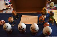 Twelve decorated eggs displayed on a table including wooden, pysanky, porcelain, and Avon eggs arranged with a lot number card.