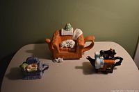 Four porcelain teapots shaped as miniature household scenes on a plain table under warm light.