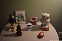 Wide view of the lot showing all items arranged on a table including owl figurine, parrot flower frog, gnome, Snoopy toothpick holder, floral plaque, small container, and lidded trinket box