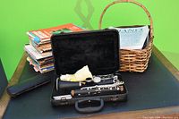 Photo showing clarinet case open with clarinet pieces and cleaning cloth inside, with sheet music stacked behind in a basket