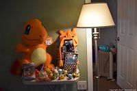 Shows a large orange Charizard plush sitting next to a small white table with multiple Pokemon figures, unopened tins, a Pez dispenser, and a Pokemon-themed hat.