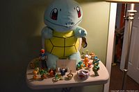 Large Squirtle plush surrounded by various Pokemon figures on a table under lamp lighting