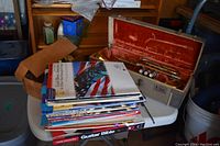 Stack of assorted music books on white plastic table, with trumpet case nearby