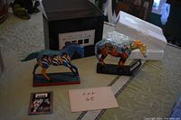 Photo showing two painted pony sculptures, one blue with riders and one yellow and blue with figures, along with packaging box labeled 'The Trail of Painted Ponies'.