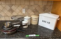 Photo of white bread bin, cookie tin, two white enamelled canisters with lids, stainless steel 750 ml pot, and two Teflon frying pans on granite counter.