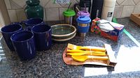 Photo of four large blue ceramic mugs, two ceramic trays stacked, a blue ceramic vase, three plastic water bottles with colored lids, four metal travel mugs with metal straws, and a box of plastic utensils with yellow cutlery visible.