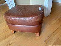 Front angled view of the medium brown leather stool showing the padded seat and wooden legs on a hardwood floor.