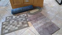 Five floor mats laid out on tile floor: three taupe rectangular mats and two braided oval mats in blue and multicolor.