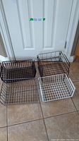 Four metal hanging racks in black, dark gray, and white finish placed on the floor in front of a door, showing wire grid construction and flat bottoms.