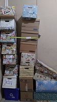 Photo shows a stacked assortment of 10 storage and file boxes with various decorative wraps including maps, holiday themes, and cartoon faces. Boxes are primarily cardboard with handle cutouts, stacked against a beige wall.