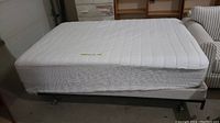 Side view of white mattress with fitted mattress pad on the metal bed frame in garage setting.