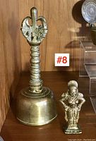 Full view of Asian brass bell and small brass figure on wooden shelf, showing size and detail.