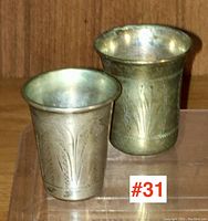 Pair of silver shot cups displayed on a wooden shelf with etched designs visible.