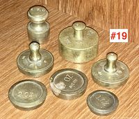 Photo showing all seven brass scale weights arranged on a wooden surface, displaying varied sizes and stamped weight markings including 5oz, 4oz, 2oz, 1oz.