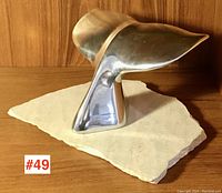 Front left angled view of the polished aluminum whale tail sculpture on natural stone base.