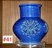 Full front view of vibrant blue art pottery vase with embossed medallion design on the body, sitting on clear stand in wood cabinet.