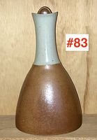 Signed Canadian art pottery carafe with brown matte glaze lower body and light blue glazed neck, with rounded ceramic stopper on top, displayed on shelf with wooden backdrop.