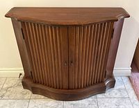 Front view of antique style pine cabinet showing round front with vertical fluted doors and two door handles. Floor tile beneath and plain wall background.