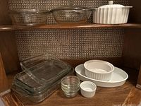 Photo showing three clear glass casserole dishes with lids, a white ceramic casserole dish with ribbed design and lid on top shelf, along with oval white ceramic baking dish, two white ceramic ramekins, and three small glass ramekins on lower shelf.