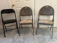 Three folding chairs: two gray metal frame with beige cushions and decorative backrest, one black metal frame with solid seat and backrest, placed on tiled floor against a door.