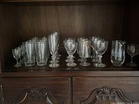 Front view of cabinet shelf displaying a variety of clear glasses arranged in rows and a pitcher on the right.