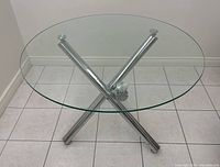 Top angled view of round glass dining table with chrome crossed tube base on tiled floor.