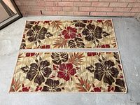 Two rectangular outdoor mats laid side-by-side on concrete, showing full floral pattern with large brown, red, and orange flowers on beige background.