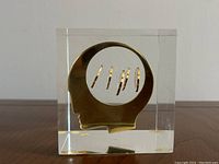 Front view showing the modernist gold-tone head silhouette with circular cutout and pennies suspended inside the cube.