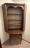Wooden vintage corner display cabinet placed against wall. Shows three shelves with woven back panel, drawer at bottom, and tapered legs.