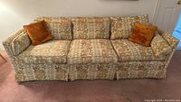 Front view of vintage floral fabric sofa with three seat cushions and two orange pillows on a carpeted floor.