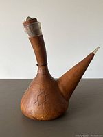 Front-left angled view showing bulbous leather body with geometric burnished pattern and wooden neck capped with metal ring and stopper.