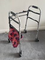 Silver-colored aluminum walker with black hand grips and a floral fabric pouch attached to the front.