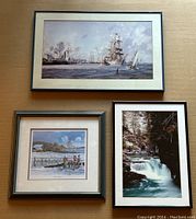 Three framed art pieces displayed on a light wood surface: top frame shows a nautical print of historic ships; bottom left frame shows a Canadiana rural scene with people near a fence; bottom right frame shows a photographic print of a waterfall in a forest.