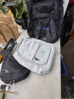 Photo showing three backpacks: a light gray Herschel, a black ZOOK, and another black backpack. Shelving and some wicker baskets in background.